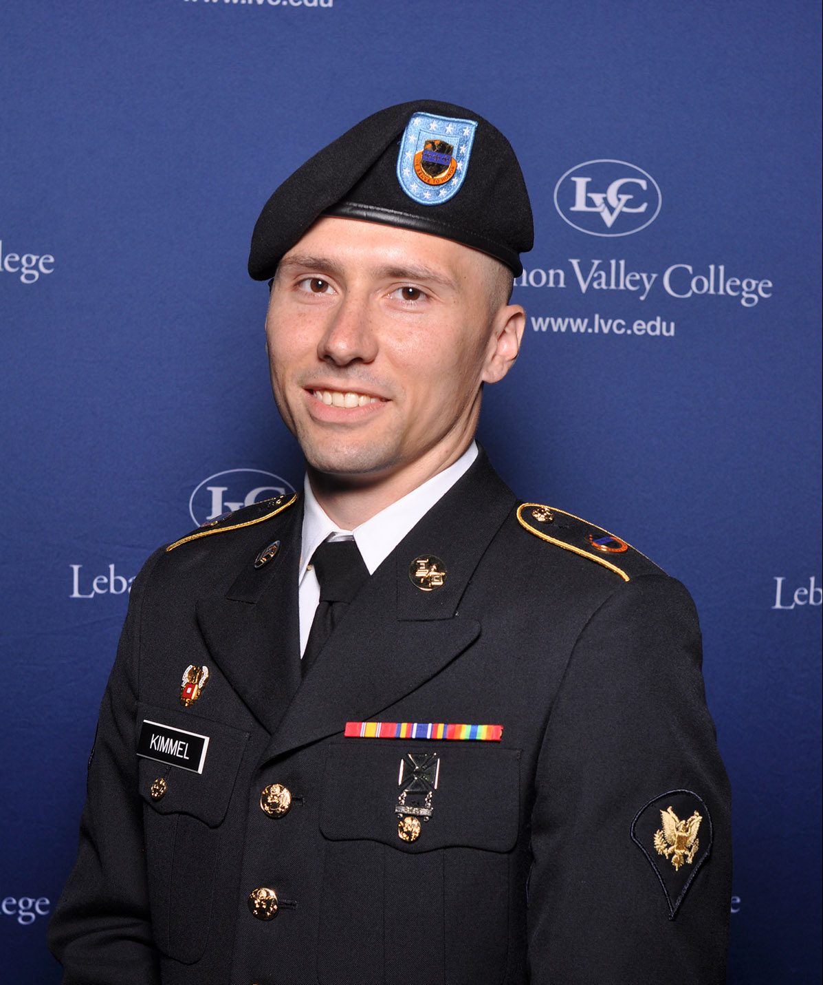 Military graduate of LVC in full uniform standing in front of LVC's logo and name on a blue wall