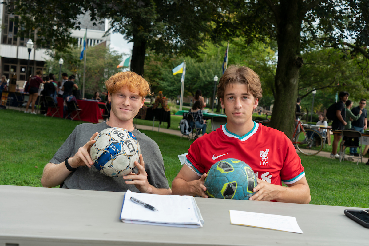 two male students sitting at engagement fair table holding soccer balls
