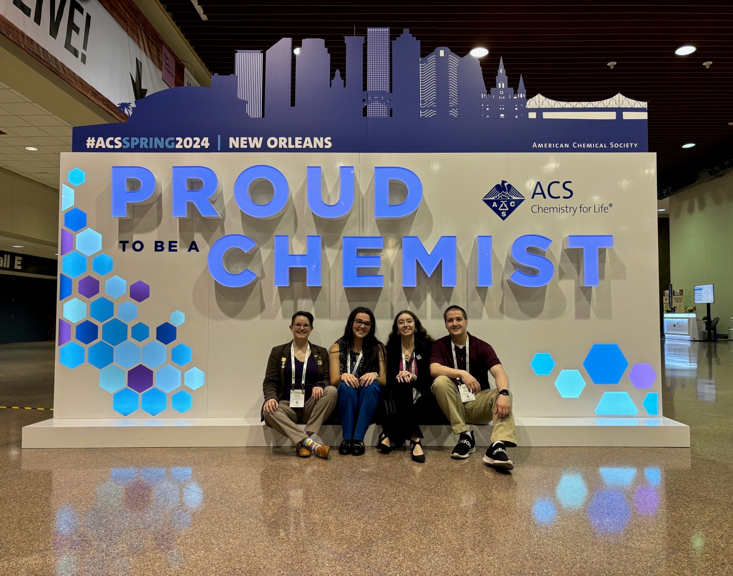 Left to right: Dr. Liz Sterner, Jacquelyn McBride ‘24, Nicole Gross ‘24, and Dylan Correll ‘24 sit in front of "Proud to be a Chemist" sign