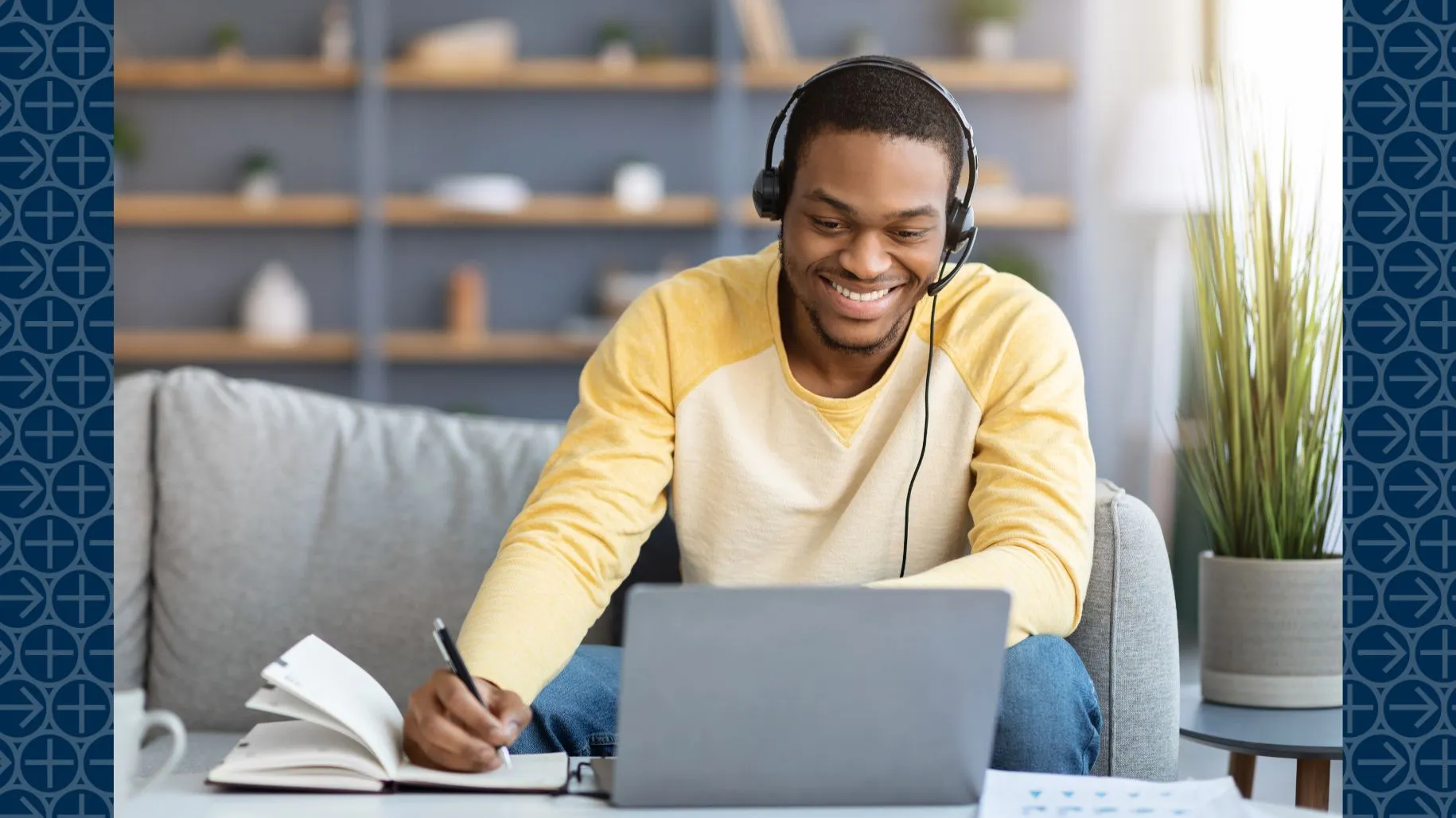 Young man wearing headset smiles at laptop screen while taking notes